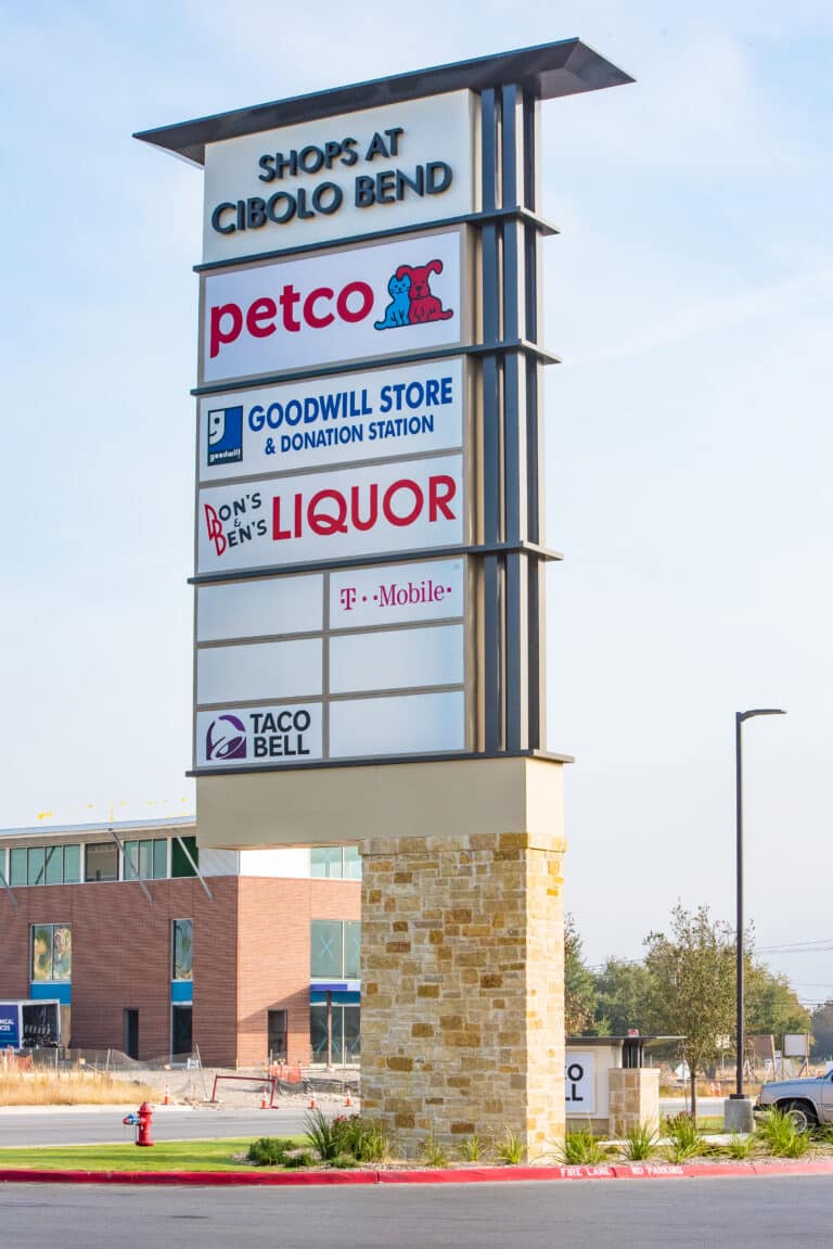A tall shopping center sign reads Shops at Cibolo Bend with logos for Petco, Goodwill Store & Donation Station, Dons & Bens Liquor, T-Mobile, and Taco Bell. A brick building and parking lot are in the background.